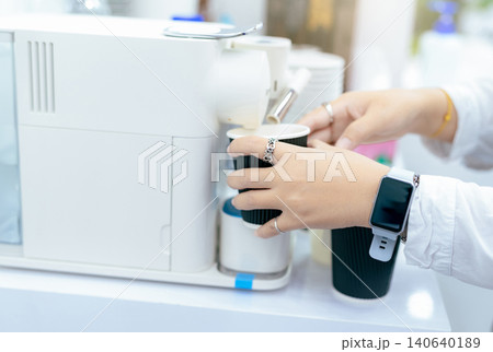 Woman making a cup of hot coffee with capsule coffee machine. Woman holding black cup taking coffee from a capsule Modern machine on the table. Espresso maker. Morning drink. International coffee day. Woman making a cup of hot coffee with capsule coffee machine. Woman holding black cup taking coffee from a capsule Modern machine on the table. Espresso maker. Morning drink. International coffee day. 140640189