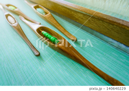 Green yarn in weaving shuttle tool on weaving machine. Textile fabric weave. Weaving using traditional weaving loom and shuttle. Textile or cloth production in Thailand. Green handmade cloth. Green yarn in weaving shuttle tool on weaving machine. Textile fabric weave. Weaving using traditional weaving loom and shuttle. Textile or cloth production in Thailand. Green handmade cloth. 140640229