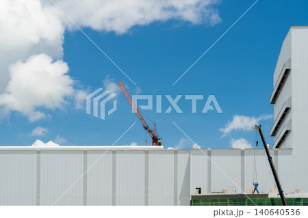 Modern industrial factory construction with crane under blue sky at manufacturing site, contemporary warehouse architecture and infrastructure development for business expansion and production growth. Modern industrial factory construction with crane under blue sky at manufacturing site, contemporary warehouse architecture and infrastructure development for business expansion and production growth. 140640536