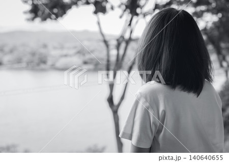 Back view of a sad Asian woman sitting alone in a park overlooking a lake. Depression, anxiety, loneliness and emotional distress in a black and white mental health. Personal wellbeing challenge. 140640655