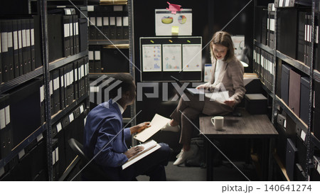 In room crammed with clues and records, Caucasian female investigator sits on table analyzing evidence with police officer, African American inspector hands over case data to policewoman with laptop. In room crammed with clues and records, Caucasian female investigator sits on table analyzing evidence with police officer, African American inspector hands over case data to policewoman with laptop. 140641274