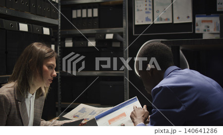 Seated on floor in incident room, African American investigator and Caucasian policewoman look for clues and evidence, female inspector with laptop, discussing criminal case with police officer. 140641286