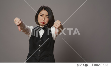 Receptionist showing thumbs down sign over grey background, expressing negativity and disapproval in studio, woman hotel concierge giving dislike symbol, feeling displeased. Receptionist showing thumbs down sign over grey background, expressing negativity and disapproval in studio, woman hotel concierge giving dislike symbol, feeling displeased. 140641528