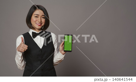 Asian hotel concierge pointing at phone with green screen layout, holding device running isolated chromakey template, receptionist showing smartphone display with copy space mockup. Asian hotel concierge pointing at phone with green screen layout, holding device running isolated chromakey template, receptionist showing smartphone display with copy space mockup. 140641544