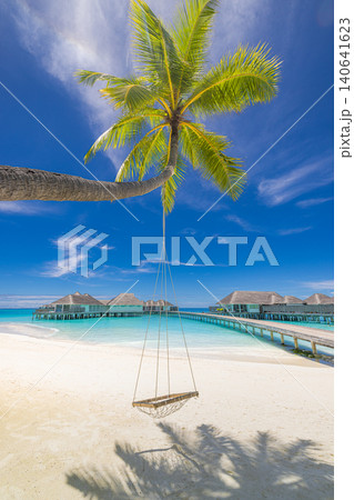 Relaxing beach swing hanging from a palm tree above turquoise water with overwater bungalows beyond. Romantic Maldives paradise scene. Stunning summer travel background with white sand and blue sky Relaxing beach swing hanging from a palm tree above turquoise water with overwater bungalows beyond. Romantic Maldives paradise scene. Stunning summer travel background with white sand and blue sky 140641623