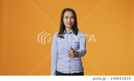 Filipino person showing thumbs up sign on camera, expressing good vibes and positivity over orange background, with young smiling woman giving like in studio, presenting approval gesture. 140641659