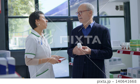Aged man in pharmacy asking pharmacist for medical opinion, needing treatment for heart ache, with qualified pharmacist helping senior client choose best pharmaceutical cure for his disease. Aged man in pharmacy asking pharmacist for medical opinion, needing treatment for heart ache, with qualified pharmacist helping senior client choose best pharmaceutical cure for his disease. 140641668