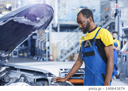 BIPOC specialist in car service using professional mechanical tool to repair broken motor, with trained specialist in garage fixing client automobile, ensuring optimal automotive performance. BIPOC specialist in car service using professional mechanical tool to repair broken motor, with trained specialist in garage fixing client automobile, ensuring optimal automotive performance. 140641674