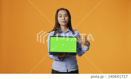 Confident adult showing laptop with green screen in studio, presenting isolated mockup screen on wireless computer, with person holding computer with chroma key display on camera. Confident adult showing laptop with green screen in studio, presenting isolated mockup screen on wireless computer, with person holding computer with chroma key display on camera. 140641727