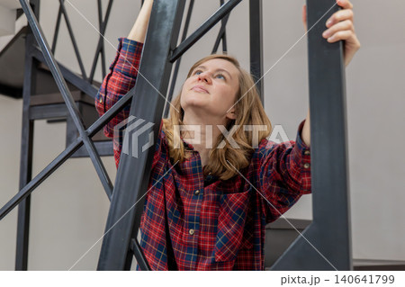 Woman dusting railing on staircase, wearing plaid shirt and jeans, standing on ladder, looking upward with focused expression, sunlight filling modern apartment interior, tidy shelves and cloth duster Woman dusting railing on staircase, wearing plaid shirt and jeans, standing on ladder, looking upward with focused expression, sunlight filling modern apartment interior, tidy shelves and cloth duster 140641799