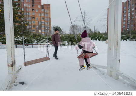 Child swinging toward camera in snow mid-air motion with visible swing chains, caregiver walking nearby in puffer coat, footprints on compacted pathway, overcast sky and apartment buildings Child swinging toward camera in snow mid-air motion with visible swing chains, caregiver walking nearby in puffer coat, footprints on compacted pathway, overcast sky and apartment buildings 140641822