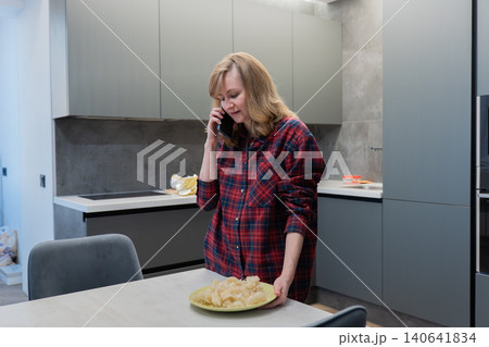 Blonde white woman talking on phone near dining table with fresh pie, multitasking between call and food prep. Plaid shirt and modern cabinetry frame a cozy but efficient kitchen scene. Expression Blonde white woman talking on phone near dining table with fresh pie, multitasking between call and food prep. Plaid shirt and modern cabinetry frame a cozy but efficient kitchen scene. Expression 140641834