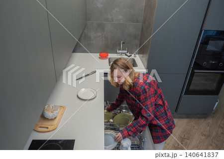 Young white woman unloading dishwasher racks in sleek kitchen after meal, placing plates into drying area while wearing cozy plaid shirt, warm indoor light, cookware and utensils nearby, relaxed Young white woman unloading dishwasher racks in sleek kitchen after meal, placing plates into drying area while wearing cozy plaid shirt, warm indoor light, cookware and utensils nearby, relaxed 140641837
