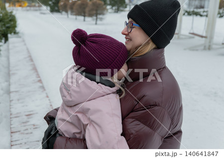 Mother carrying sleepy daughter in arms on snowy sidewalk, close forehead contact and gentle kiss, layered coats and warm beanies, calm urban morning mood, visible breath and muted color palette Mother carrying sleepy daughter in arms on snowy sidewalk, close forehead contact and gentle kiss, layered coats and warm beanies, calm urban morning mood, visible breath and muted color palette 140641847