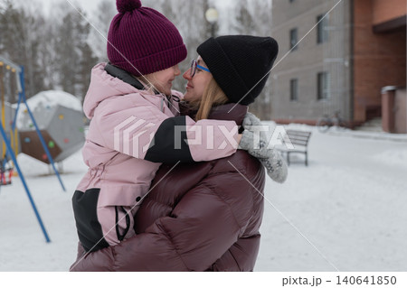 white aunt kissing daughters nose under snowy sky, tender forehead and cheek contact, warm puffer coats and mittens, playful grip on climbing ropes, playground structure and building behind, 140641850