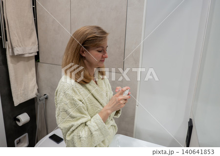 Woman in bathrobe applying serum in bathroom mirror, beauty blogger testing new skincare product at vanity, glass bottle held near face, soft morning light, neutral tiles and folded towel, focused 140641853