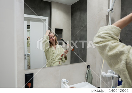 Caucasian woman at mirror brushing hair in bathrobe, concentrated expression, practicing poses and styling techniques, modern sink and vanity visible, prepping for studio photoshoot with poised 140642002
