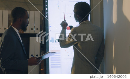 Supervisor showing employee useful datasets on digital screen to help him finish project tasks, with business executives looking over accounting figures in office during nightshift. 140642003