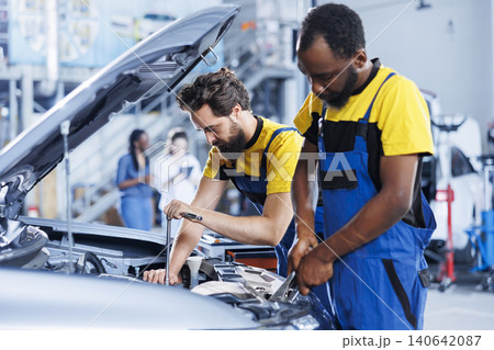 Certified servicemen in auto repair shop working together on fixing car, discussing best options, with bIPOC worker and colleague collaborating on servicing broken vehicle. 140642087