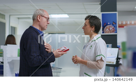Certified pharmacist in pharmacy offering patient heart arrhythmia medication antibiotic to treat ailment symptoms, with older man receiving heart attack prevention tablets from wellness speicalist. Certified pharmacist in pharmacy offering patient heart arrhythmia medication antibiotic to treat ailment symptoms, with older man receiving heart attack prevention tablets from wellness speicalist. 140642149