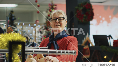 Elderly woman browsing through clothes in Christmas decorated clothing store during winter holiday season, with aged customer at the mall in shopping spree session. Elderly woman browsing through clothes in Christmas decorated clothing store during winter holiday season, with aged customer at the mall in shopping spree session. 140642246