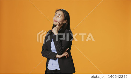 Thoughtful businesswoman posing over orange backdrop, thinking about new ideas and being in doubt, with clueless uncertain person brainstorming solutions, acting focused in front of camera. Thoughtful businesswoman posing over orange backdrop, thinking about new ideas and being in doubt, with clueless uncertain person brainstorming solutions, acting focused in front of camera. 140642253