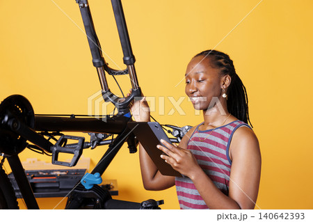 Young woman inspecting damaged bicycle and exploring repair options on her smart tablet, with image of woman checking broken bike components while holding a digital device. 140642393