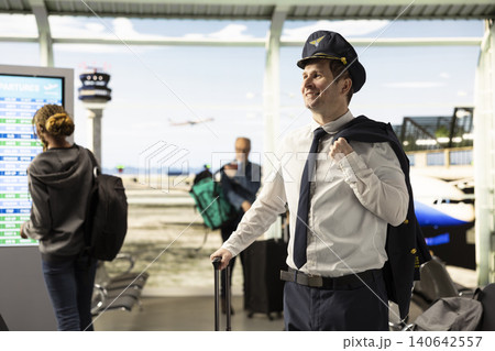 Airline captain in uniform walking through busy airport terminal with suitcase, preparing for flight. Professional pilot embody aviation career, reliable service within cabin crew operations. Airline captain in uniform walking through busy airport terminal with suitcase, preparing for flight. Professional pilot embody aviation career, reliable service within cabin crew operations. 140642557