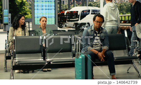 Black solo traveler waiting in busy coach station before intercity departure, sitting with baggage and backpack while reviewing schedule and departure time. Long-haul highway bus travel 140642739