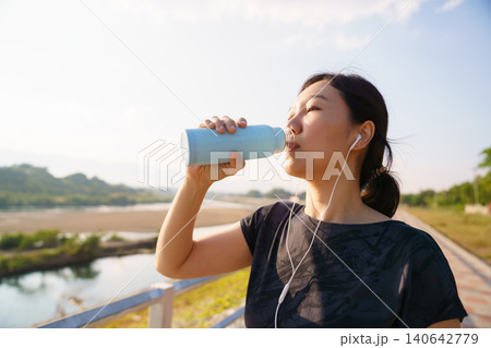 Asian woman drinking water after outdoor workout with earphones Asian woman drinking water after outdoor workout with earphones 140642779