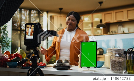 Vertical chroma key on tablet display with food vlogger filming cooking process, behind the scenes with woman influencer making a recipe at kitchen counter, home studio with meal prep. 140642788