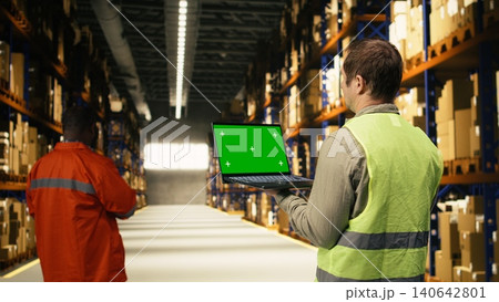 Green screen on laptop held by male worker in fulfillment center depot, operational workforce handling freight and packages prepared for distribution. E-commerce shipment operations. Green screen on laptop held by male worker in fulfillment center depot, operational workforce handling freight and packages prepared for distribution. E-commerce shipment operations. 140642801