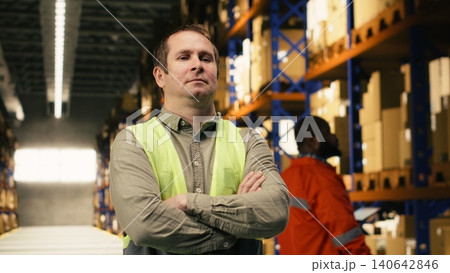 Portrait of adult employee in a logistics fulfillment center near shelves, male wearing safety vest and managing packages for order fulfillment. Global e-commerce shipping operations. Portrait of adult employee in a logistics fulfillment center near shelves, male wearing safety vest and managing packages for order fulfillment. Global e-commerce shipping operations. 140642846