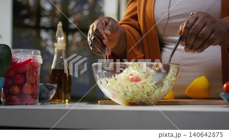 Close up of african american woman mixing vegetables in a salad bowl at home, recording online food vlog for vegetarians or a healthy eating choice. Culinary tutorial with meal prep. Close up of african american woman mixing vegetables in a salad bowl at home, recording online food vlog for vegetarians or a healthy eating choice. Culinary tutorial with meal prep. 140642875