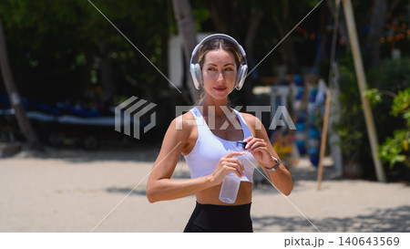 Beautiful sporty woman with headphones holding water bottle on the beach Beautiful sporty woman with headphones holding water bottle on the beach 140643569