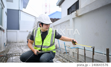 Construction Inspector Measuring Wall with Tape Measure and Tablet at Building Site 140643576