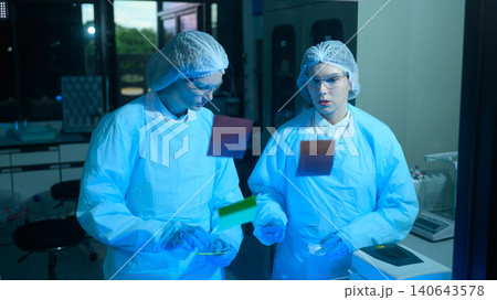 Two Female Scientists in Cleanroom Suits Examining Samples Under Blue UV Light in Modern Laboratory Two Female Scientists in Cleanroom Suits Examining Samples Under Blue UV Light in Modern Laboratory 140643578