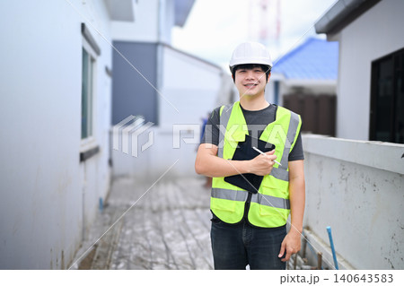 Young Asian Male Construction Inspector Smiling at Building Site Wearing Safety Vest and Hard Hat with Clipboard Young Asian Male Construction Inspector Smiling at Building Site Wearing Safety Vest and Hard Hat with Clipboard 140643583