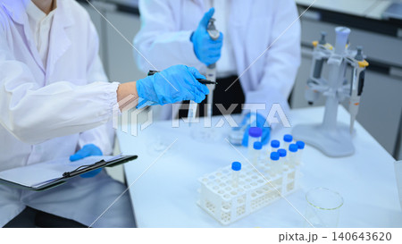 Scientists in White Lab Coats and Blue Gloves Conducting Laboratory Experiment with Test Tubes and Vials Scientists in White Lab Coats and Blue Gloves Conducting Laboratory Experiment with Test Tubes and Vials 140643620