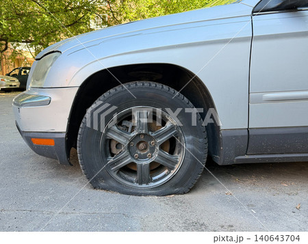 Flat tire on parked silver car stands on urban street, vehicle maintenance issue, transportation disruption, roadside safety. Automotive service, insurance communication, repair, roadside assistance Flat tire on parked silver car stands on urban street, vehicle maintenance issue, transportation disruption, roadside safety. Automotive service, insurance communication, repair, roadside assistance 140643704
