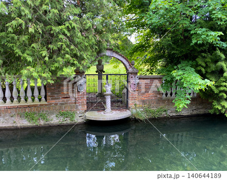 Mysterious vintage garden entrance with brick fence, statues and lush green trees by the water canal 140644189