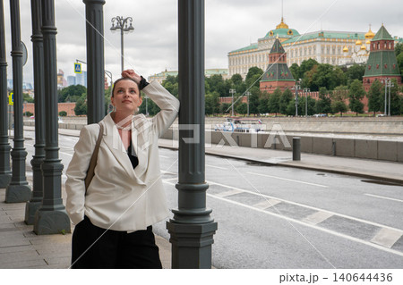 Woman stands on Sofiyskaya Embankment in Moscow with Kremlin across river, framed by colonnade and overcast sky in contemporary urban setting. Travel destination, city architecture, cultural heritage Woman stands on Sofiyskaya Embankment in Moscow with Kremlin across river, framed by colonnade and overcast sky in contemporary urban setting. Travel destination, city architecture, cultural heritage 140644436