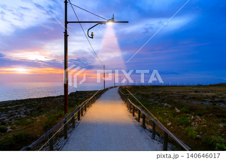 A scenic coastal walkway bathed in the warm glow of lanterns as the sun begins to rise over the tranquil sea 140646017
