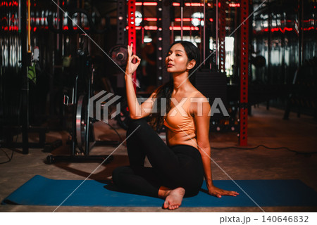 Woman seated on a blue yoga mat making a mindful hand gesture in a dark gym, representing meditation, breathing, relaxation, wellness, inner balance, calm focus, and fitness recovery. Woman seated on a blue yoga mat making a mindful hand gesture in a dark gym, representing meditation, breathing, relaxation, wellness, inner balance, calm focus, and fitness recovery. 140646832