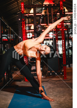 Woman holding triangle yoga pose on a blue mat in a dark gym, emphasizing flexibility, balance, side stretch, core strength, body alignment, mindful movement, wellness, and fitness routine. Woman holding triangle yoga pose on a blue mat in a dark gym, emphasizing flexibility, balance, side stretch, core strength, body alignment, mindful movement, wellness, and fitness routine. 140646834