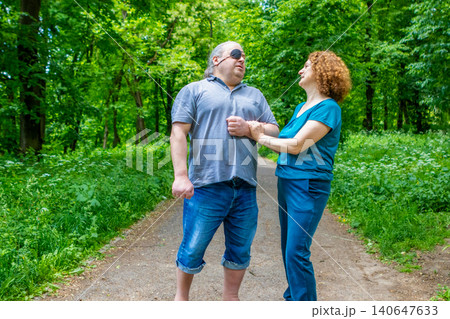 Mature couple walking in park as woman supports man wearing an eye patch during stroll Mature couple walking in park as woman supports man wearing an eye patch during stroll 140647633