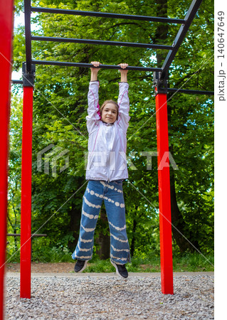 Girl hanging from monkey bars at outdoor playground practicing strength and play in green park 140647649