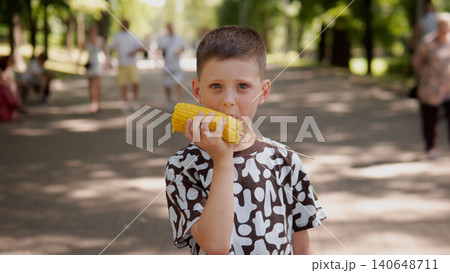 A young boy wearing a patterned shirt stands in a bright park, eating corn on the cob A young boy wearing a patterned shirt stands in a bright park, eating corn on the cob 140648711