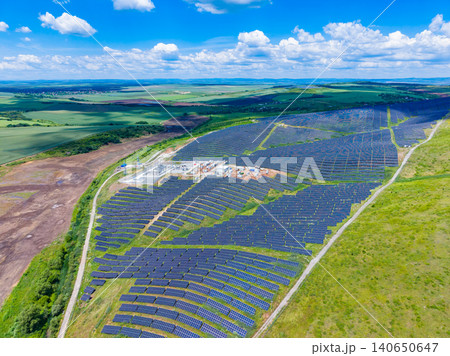 Solar farm landscape. Renewable energy installation across hilly terrain. Sunlit renewable energy farm with panels following rolling landscape contours and scattered clouds 140650647
