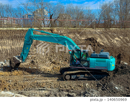 Excavator digging in a construction site near a city on a sunny day with blue sky and trees in the background 140650969